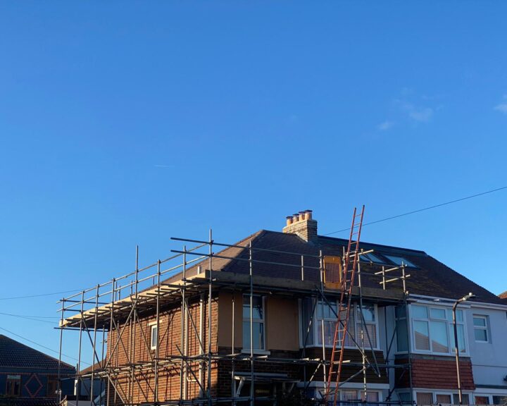 A house under construction with scaffolding and roofers at work on the flat roof.