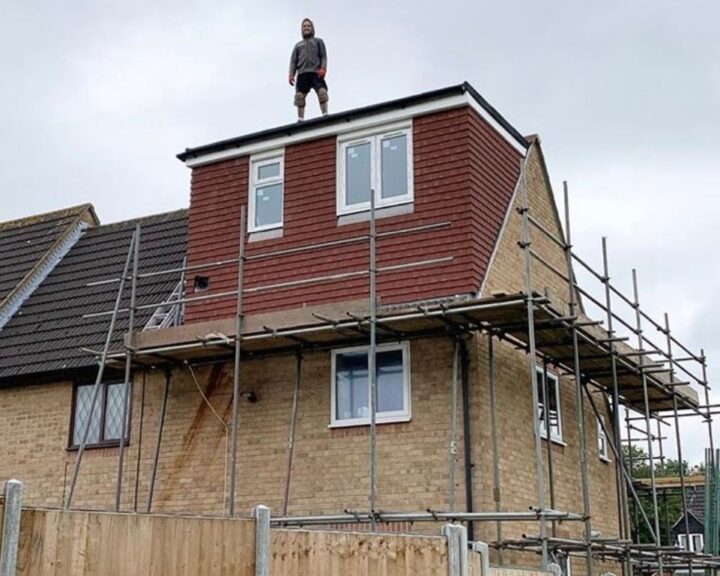 A roofer standing on top of a house with scaffolding, working on a flat roof.
